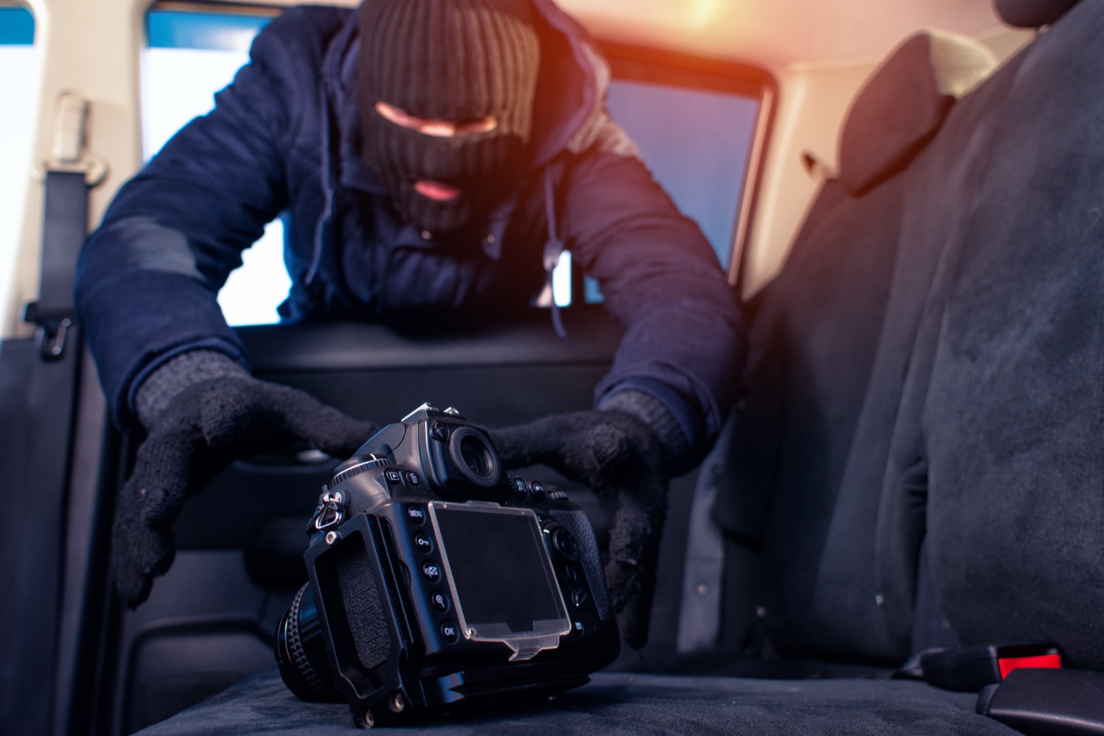 Masked thief reaching through a car window to steal a DSLR camera off the seat